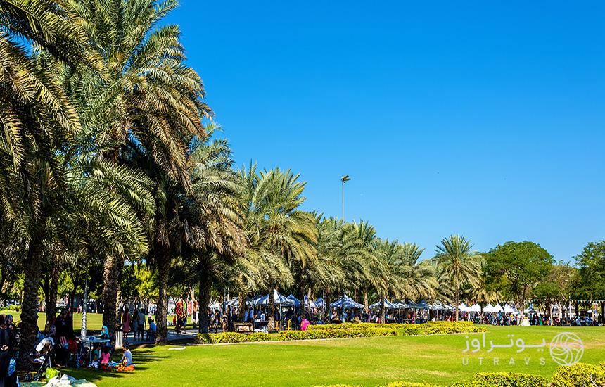 نمایی از پارک زعبیل دبی که چمن، ردیف درختان نخل و گروهی از مردم نشسته روی زمین را نشان می‌دهد.